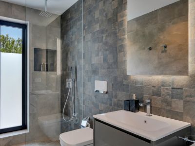 Moderno baño con ducha de lluvia y mampara de cristal. Paredes de azulejos texturizados, ventana esmerilada y tocador con lavabo integrado. Espejo retroiluminado.