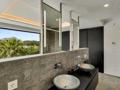 Moderno baño con lavabos dobles de cristal, salpicadero de azulejos hexagonales grises texturizados y espejos suspendidos iluminados. Amplia ventana con vistas a la montaña.