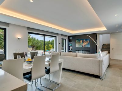 Salón-comedor de planta abierta con mesa blanca moderna, sofá gris claro y pared multimedia con chimenea eléctrica. Grandes puertas de cristal conectan con el jardín.