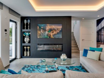 Salón moderno con pared decorativa oscura, TV integrada y chimenea eléctrica. Grandes puertas de cristal aportan luz natural y acceso al exterior, complementado por iluminación de techo.