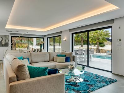 Salón moderno con sofá en L, alfombra turquesa y amplios ventanales que ofrecen acceso directo a la piscina y vistas a las palmeras.