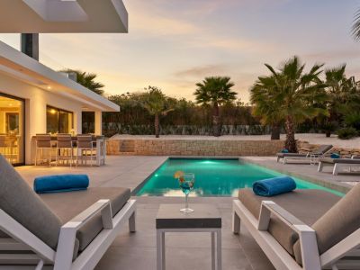 Terraza exterior con piscina iluminada al atardecer, rodeada de palmeras y una zona de bar exterior visible a través de puertas correderas de cristal.
