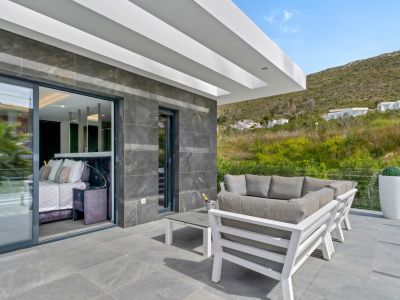 Terraza exterior con suelo de gres gris y elegante mobiliario de salón blanco. Grandes puertas correderas conectan con un dormitorio interior, ofreciendo vistas a la colina verde.
