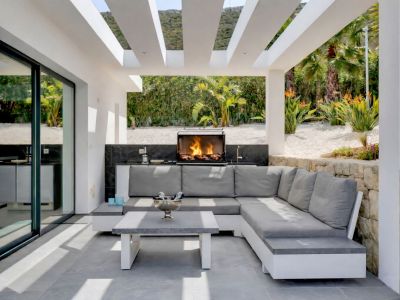 Terraza exterior cubierta con cocina integrada y barbacoa encendida, equipada con un sofá en L y mesa de centro sobre suelo de gres gris, frente a un jardín paisajístico.