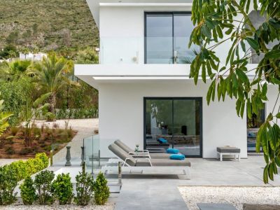 Terraza moderna con tumbonas grises sobre baldosas de gran formato, delimitada por barandillas de cristal y un jardín paisajístico. Fachada blanca con puertas correderas de cristal y fondo montañoso.