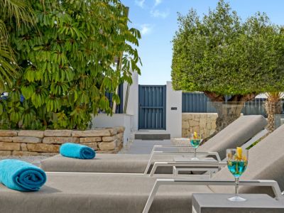 Tumbonas modernas con toallas azules y cócteles refrescantes, en un exuberante jardín mediterráneo con muro de piedra y portón azul.