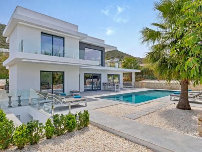 Villa blanca moderna con amplios balcones de cristal y piscina rectangular. El exterior cuenta con patio de baldosas grises, paisajismo de grava, comedor exterior y tumbonas, con fondo montañoso.