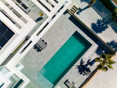Vista aérea de la moderna villa con piscina rectangular, amplias terrazas de gres gris, paneles solares en el tejado y un jardín de grava con palmeras.