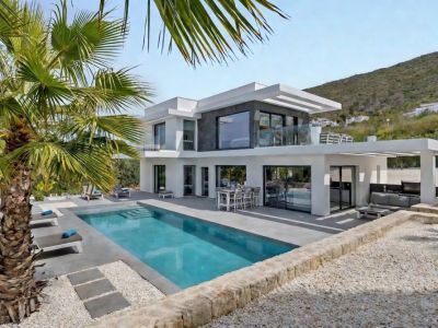 Vista exterior de la villa moderna, con su piscina rectangular, amplia terraza pavimentada con tumbonas y dos áreas de estar exteriores cubiertas, enmarcada por una palmera y una ladera natural.