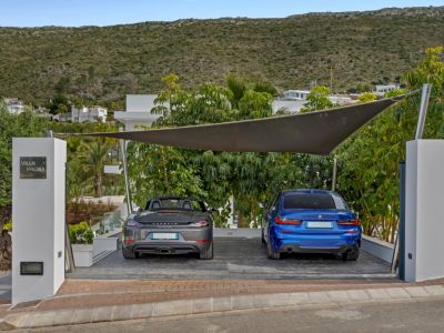 Zona de aparcamiento exterior con vela de sombra moderna, para dos coches de lujo: un Porsche gris y un BMW azul. El pilar de entrada muestra el letrero 'Villa Palma', rodeado de vegetación mediterránea y una colina al fondo.