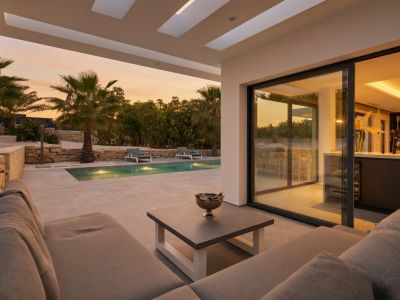 Zona de estar exterior cubierta con un sofá seccional gris moderno y mesa de centro, ofreciendo acceso directo a una piscina iluminada y jardín exuberante bajo un cielo de atardecer vibrante.