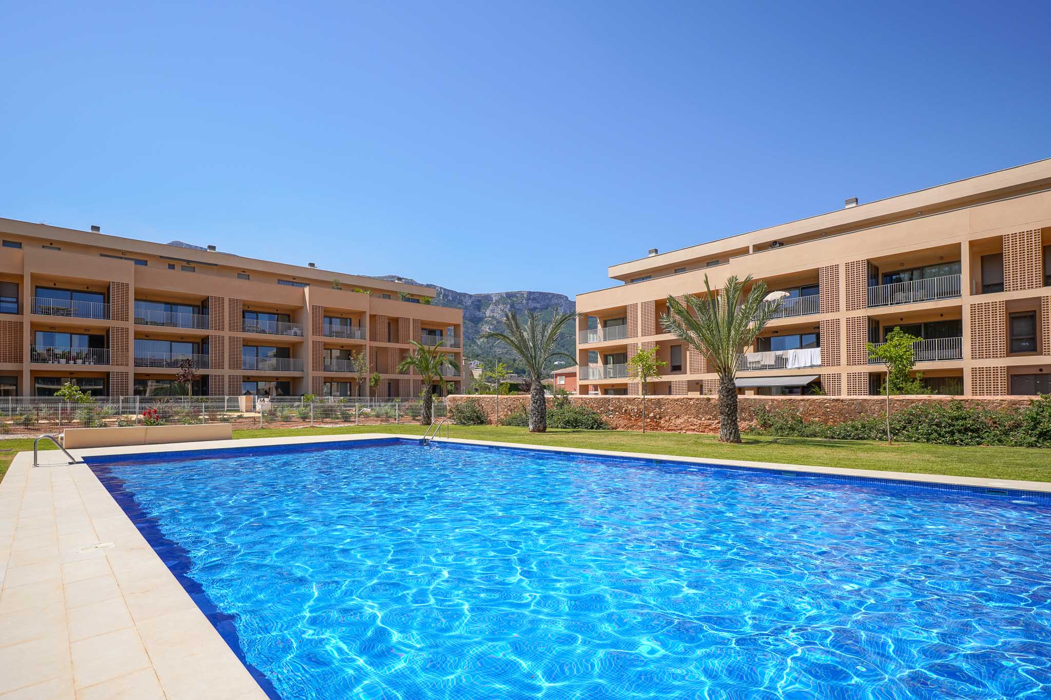 Amplia piscina comunitaria con mosaicos azules, rodeada de césped y edificios residenciales modernos con balcones. Al fondo, se aprecian palmeras y montañas.