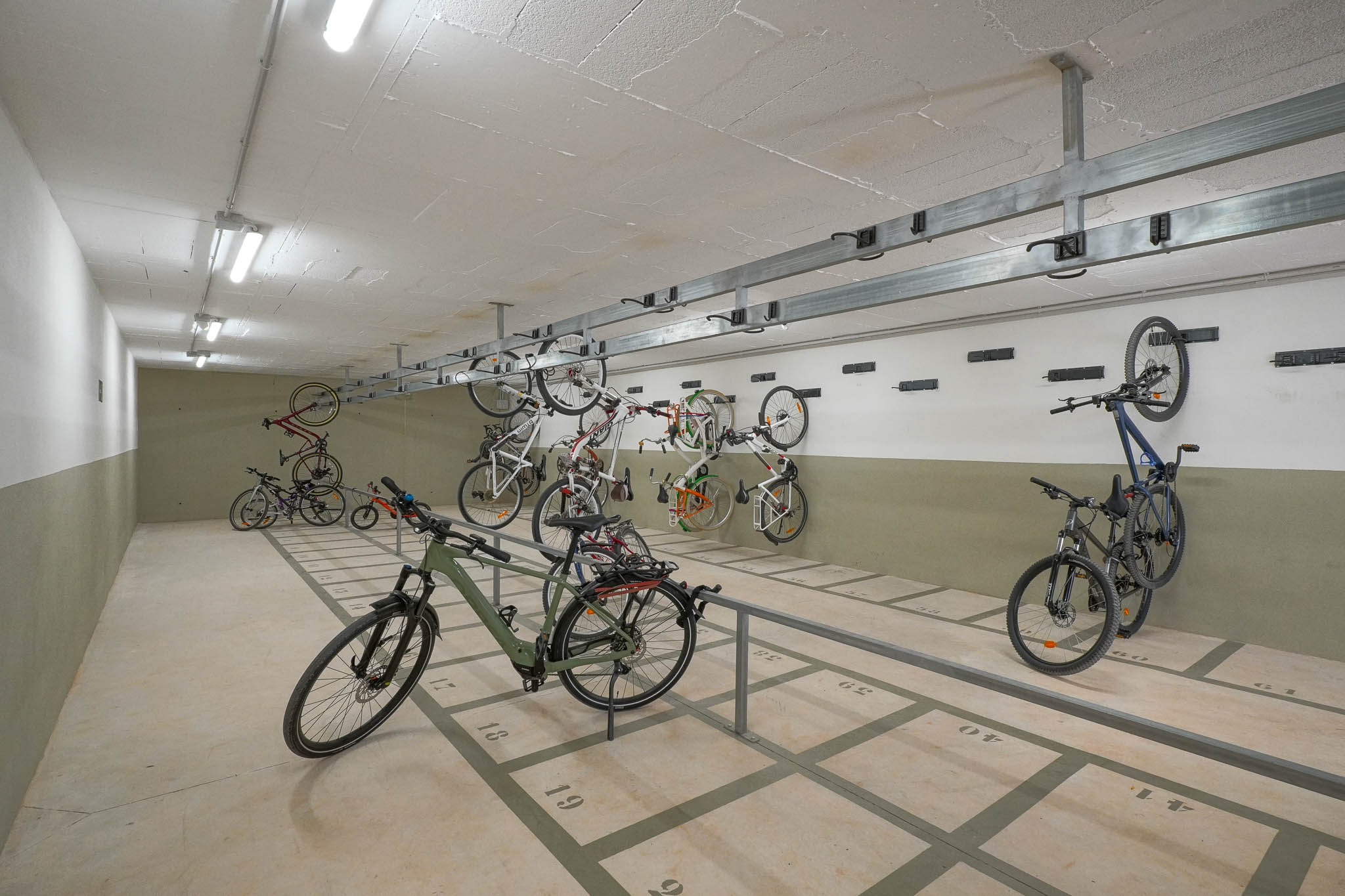 Amplia zona de aparcamiento comunitario para bicicletas con plazas numeradas en el suelo y soportes de pared. Iluminado con luces fluorescentes, ofrece almacenamiento seguro en el complejo residencial.