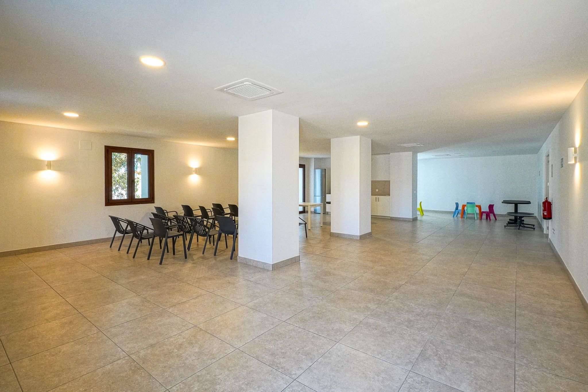Amplio centro social con suelo cerámico, pilares estructurales y luminarias empotradas. Incluye zona de sillas para reuniones y un rincón infantil con mobiliario colorido.
