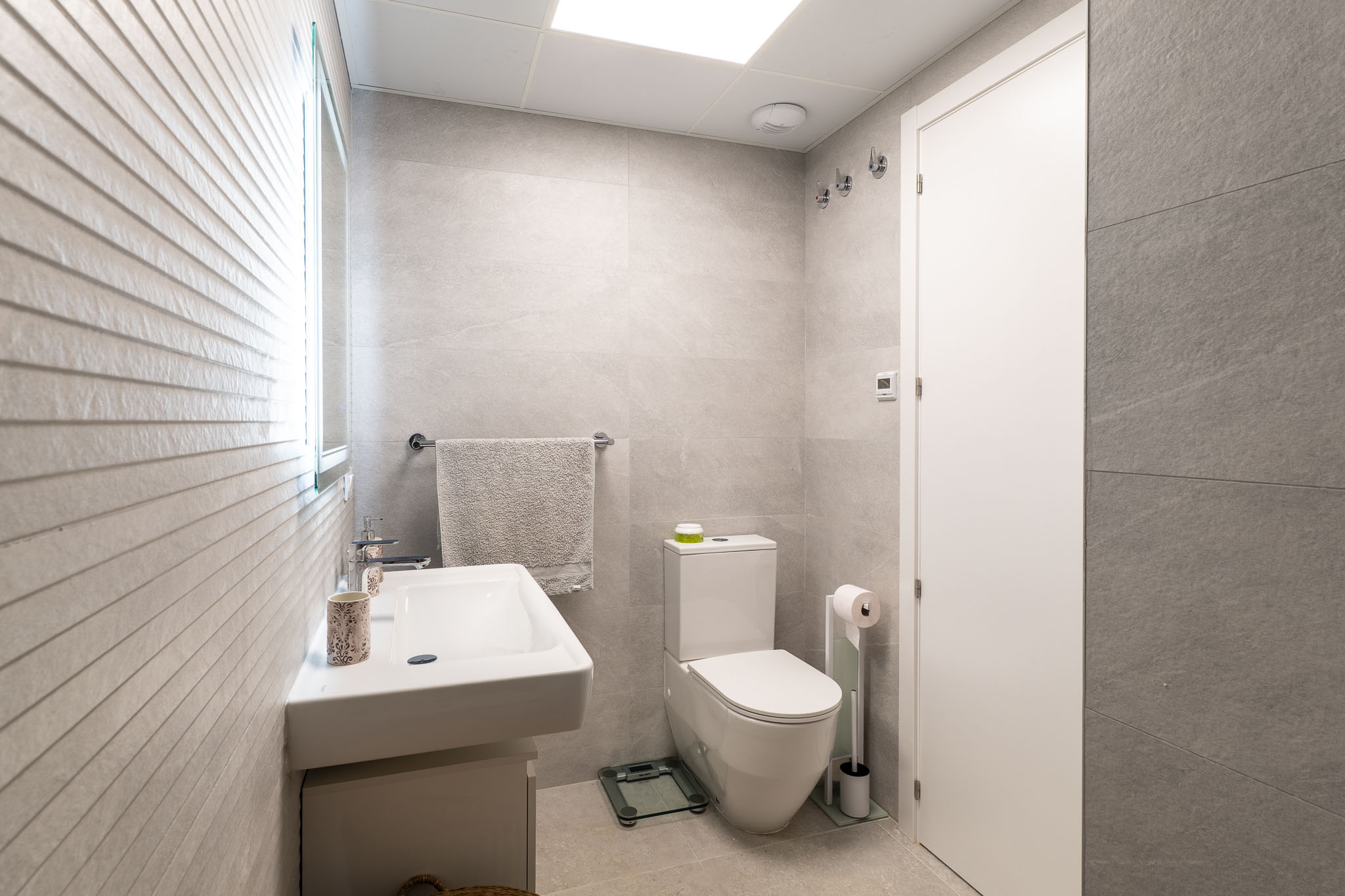 Baño moderno con azulejos grises de gran formato, lavabo suspendido y espejo iluminado. Una pared de acento presenta un diseño texturizado horizontal.