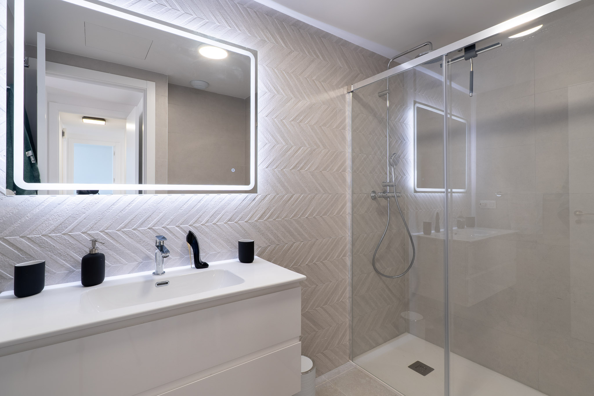 Baño moderno con lavabo blanco, espejo retroiluminado y azulejos en patrón espiga. La ducha con mampara de cristal y grifería cromada complementa el diseño elegante.