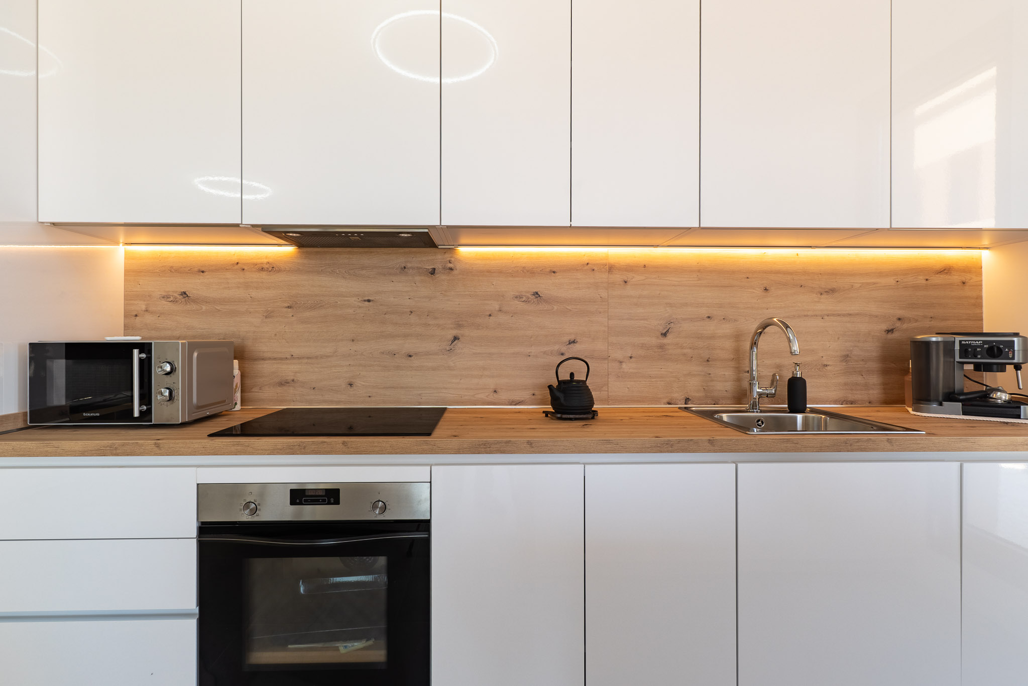 Cocina moderna con armarios blancos brillantes, encimera y salpicadero de efecto madera, e iluminación LED bajo los gabinetes. Equipada con horno, microondas y placa de inducción.