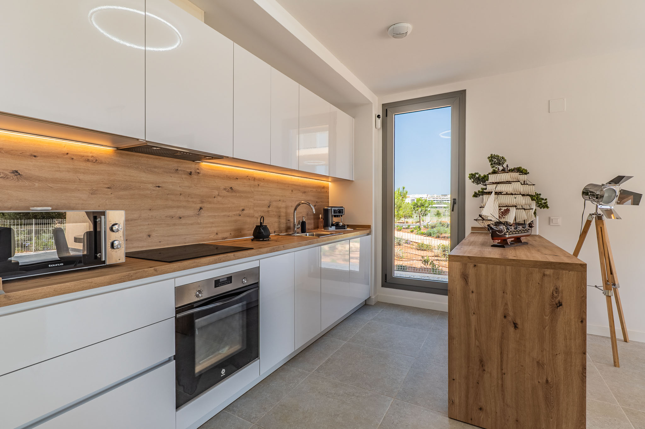 Cocina moderna con armarios blancos brillantes, encimera y salpicadero efecto madera. Equipada con electrodomésticos integrados, iluminación LED y ventana con vistas al exterior.