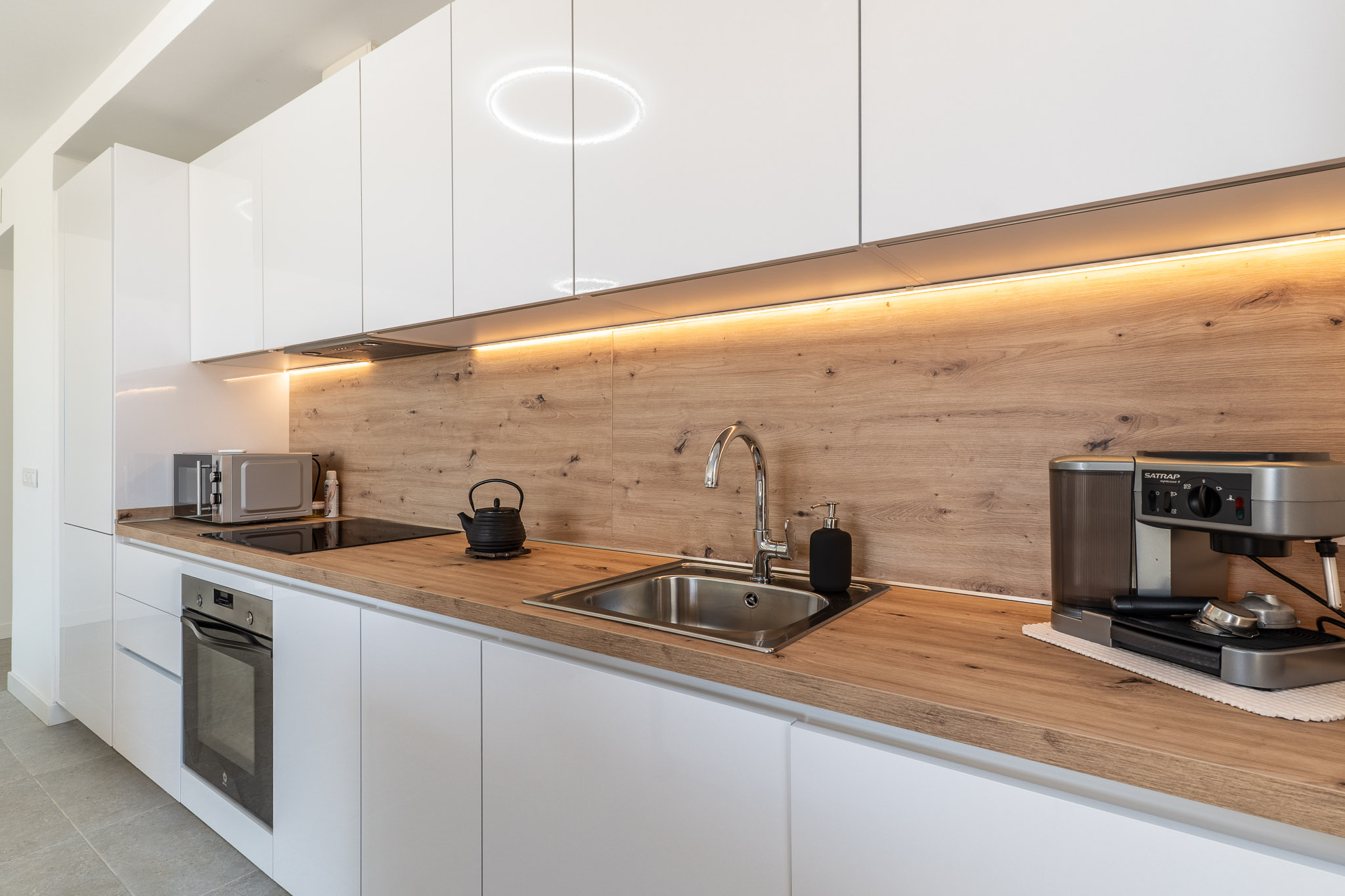 Cocina moderna con mobiliario blanco brillante, encimera y salpicadero de madera clara. Equipada con horno integrado, placa de inducción y fregadero de acero, realzada por iluminación LED bajo los armarios.