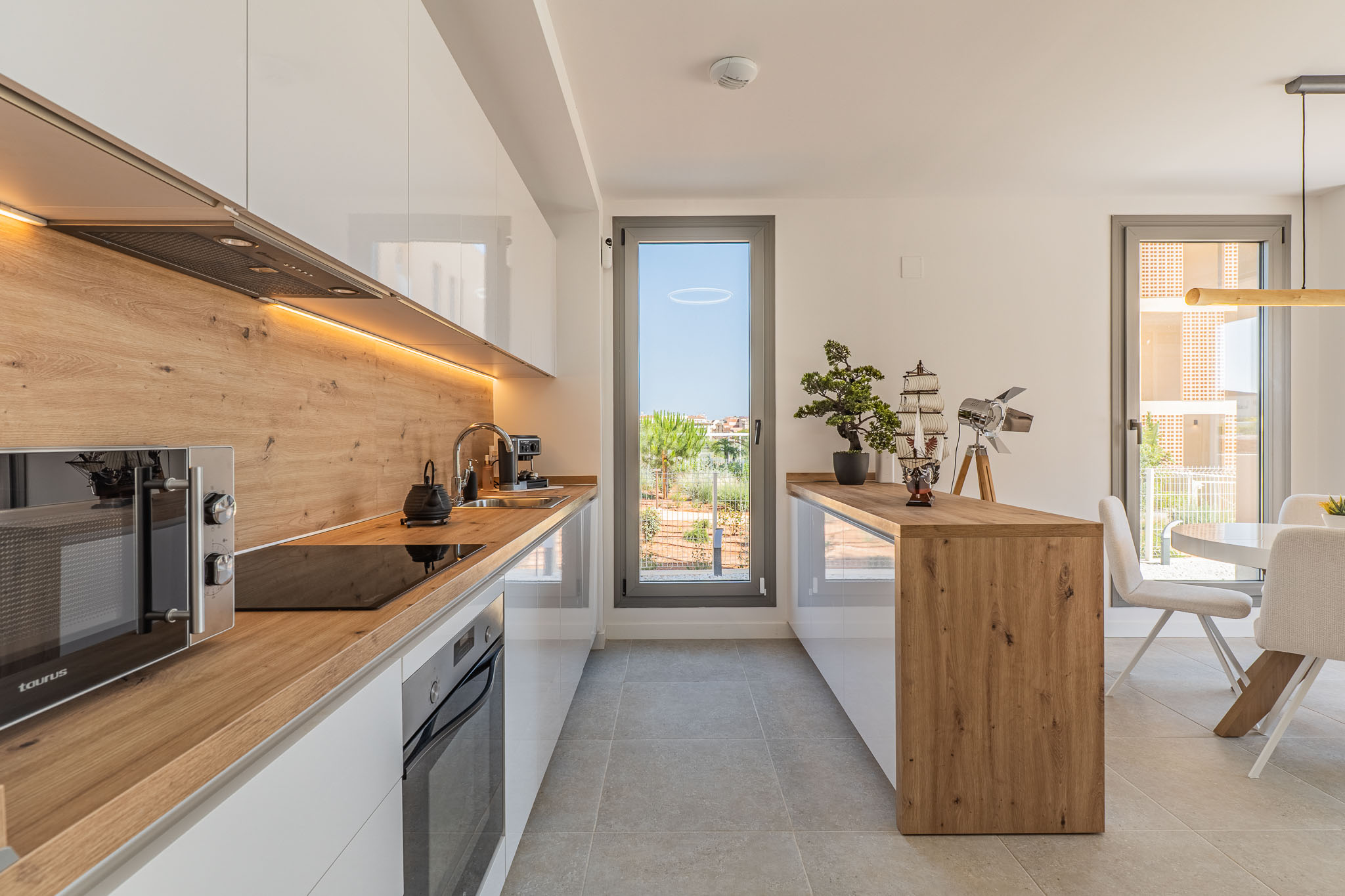 Cocina moderna de diseño abierto con muebles blancos brillantes, encimeras y salpicadero de efecto madera, e iluminación LED bajo los armarios. Incluye electrodomésticos integrados y acceso a la terraza.