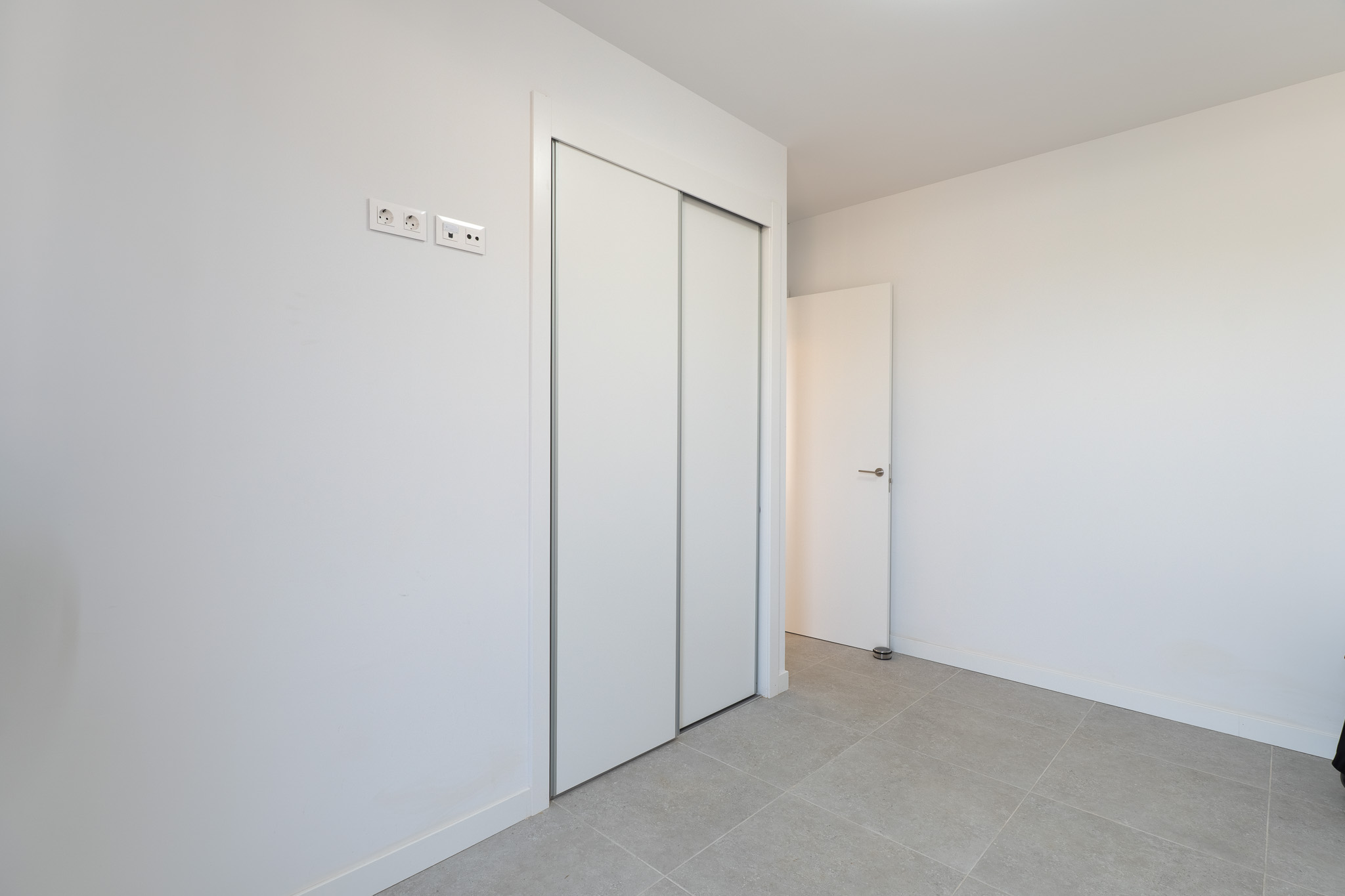 Dormitorio con amplio armario empotrado de puertas correderas blancas y suelo de gres claro. Incluye tomas eléctricas y de datos modernas.