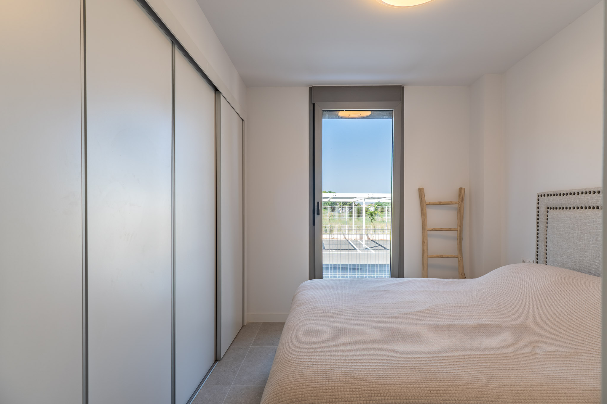 Dormitorio luminoso con armario empotrado de puertas correderas blancas, suelo porcelánico y acceso directo al exterior a través de una puerta acristalada. Cama con cabecero tapizado.