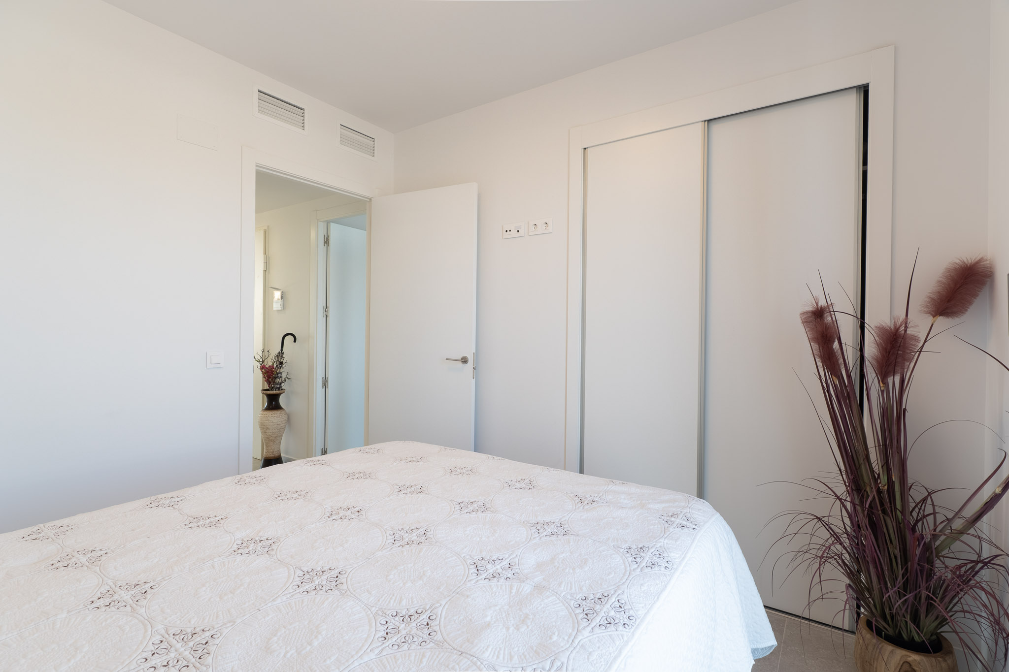 Dormitorio luminoso con cama de edredón blanco y detalles bordados. Incluye armario empotrado con puertas correderas blancas y climatización por conductos. Un toque decorativo con planta ornamental.