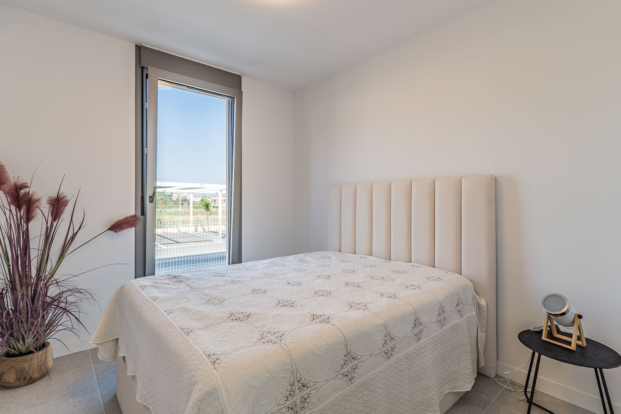 Dormitorio luminoso con cama y cabecero acolchado beige. Gran ventanal de suelo a techo con vistas exteriores y suelo de gres gris. Planta decorativa y mesita auxiliar moderna.