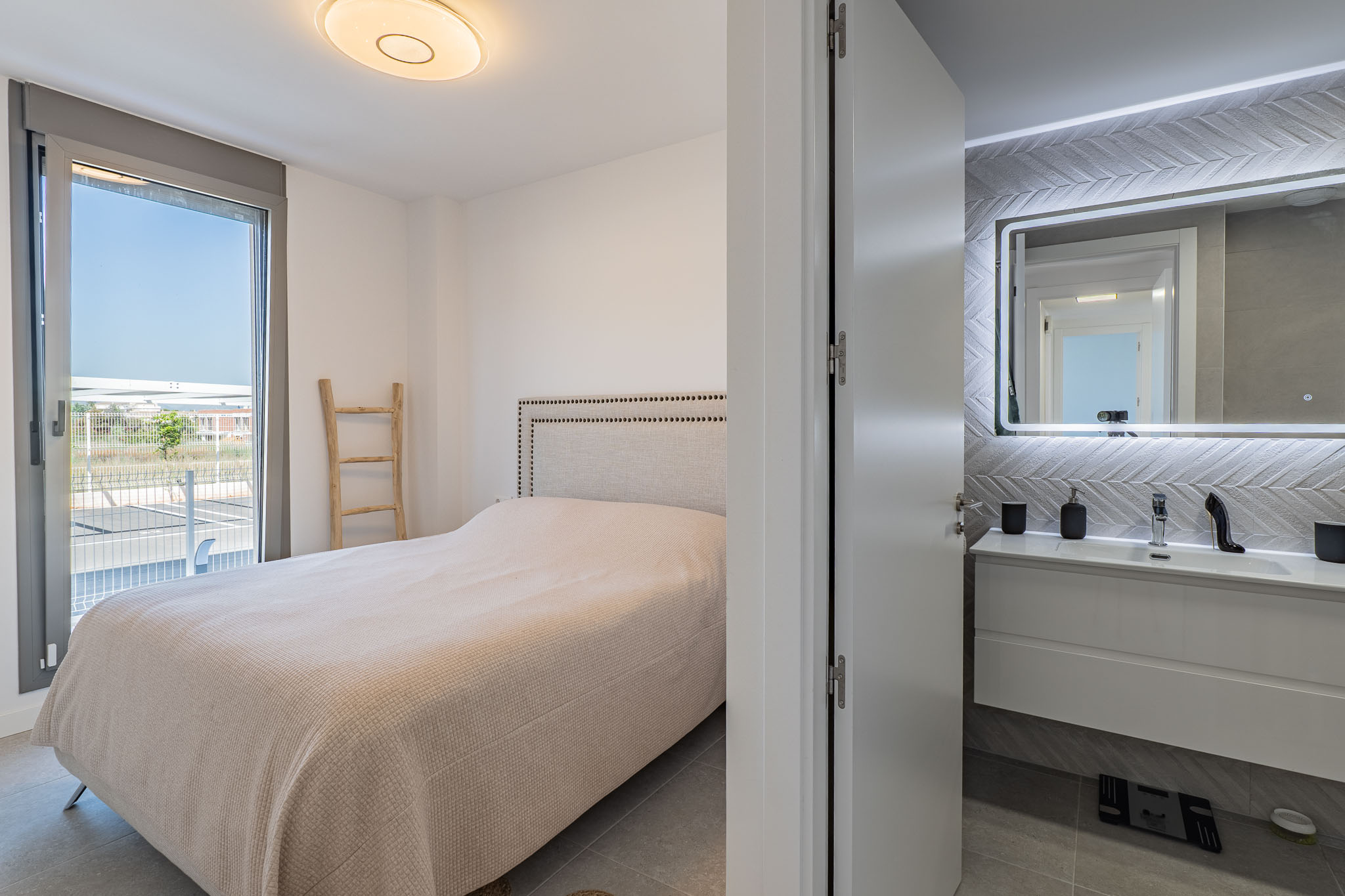 Dormitorio principal con cama de cabecero tapizado y salida directa a una terraza exterior. Se aprecia un baño en suite con lavabo flotante y espejo retroiluminado.