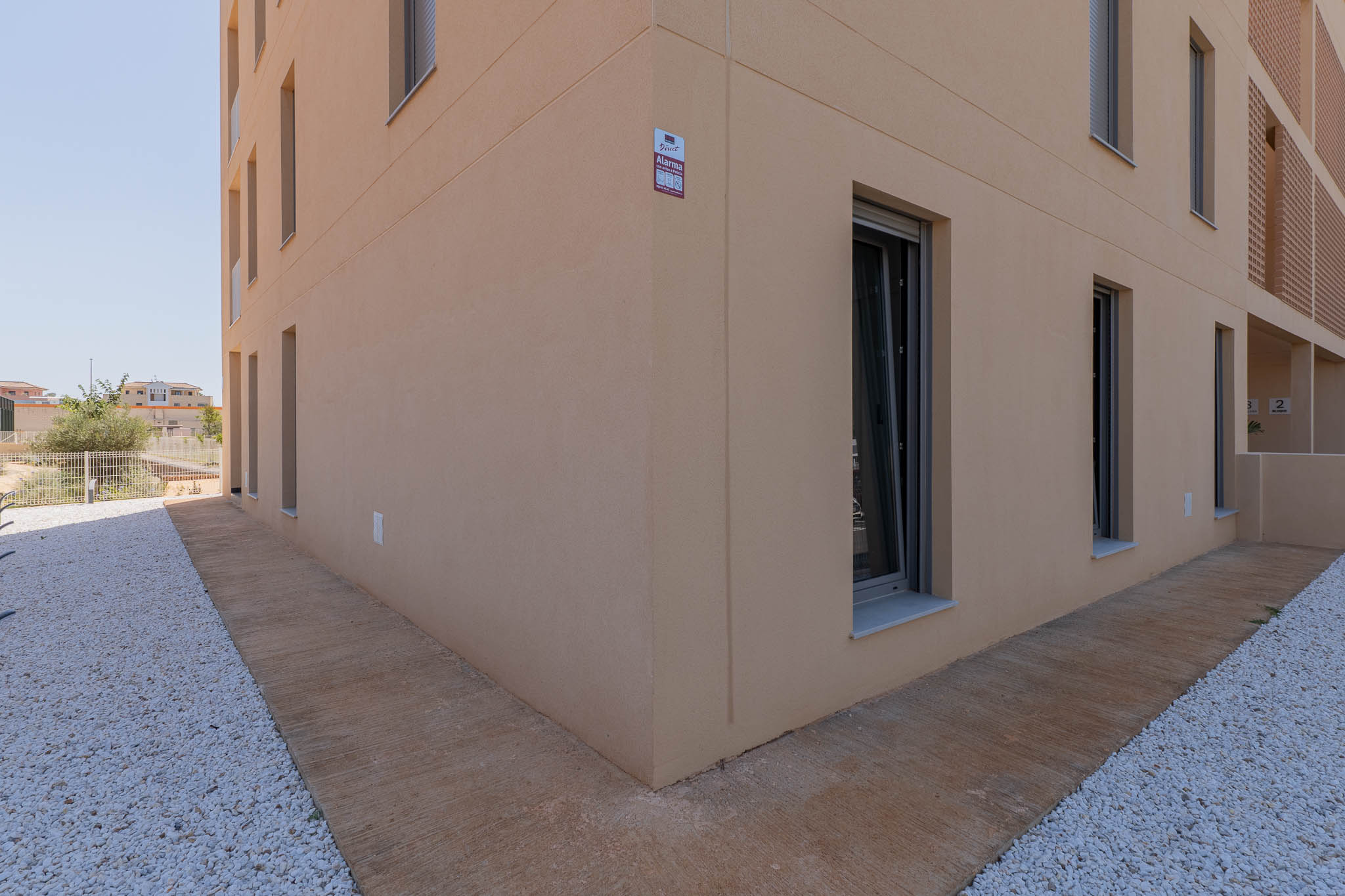 Exterior de edificio moderno con fachada de estuco beige claro y ventanas de marcos oscuros. El suelo presenta un camino de hormigón texturizado junto a un área de gravilla blanca, con alarma visible.