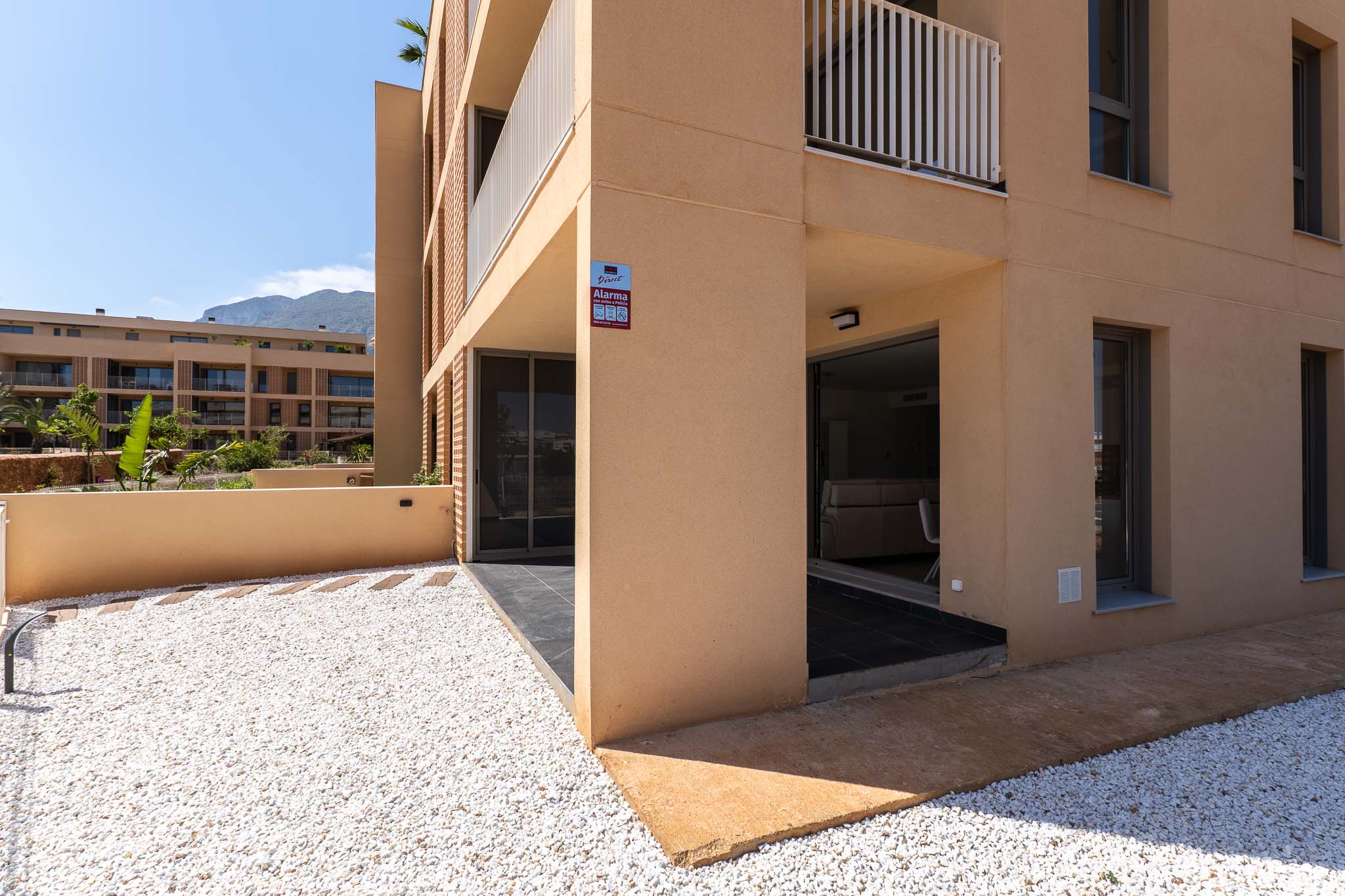 Exterior de planta baja con fachada moderna color ocre. Destaca un jardín privado de grava blanca y terraza cubierta con acceso directo al interior, con vistas a la montaña.