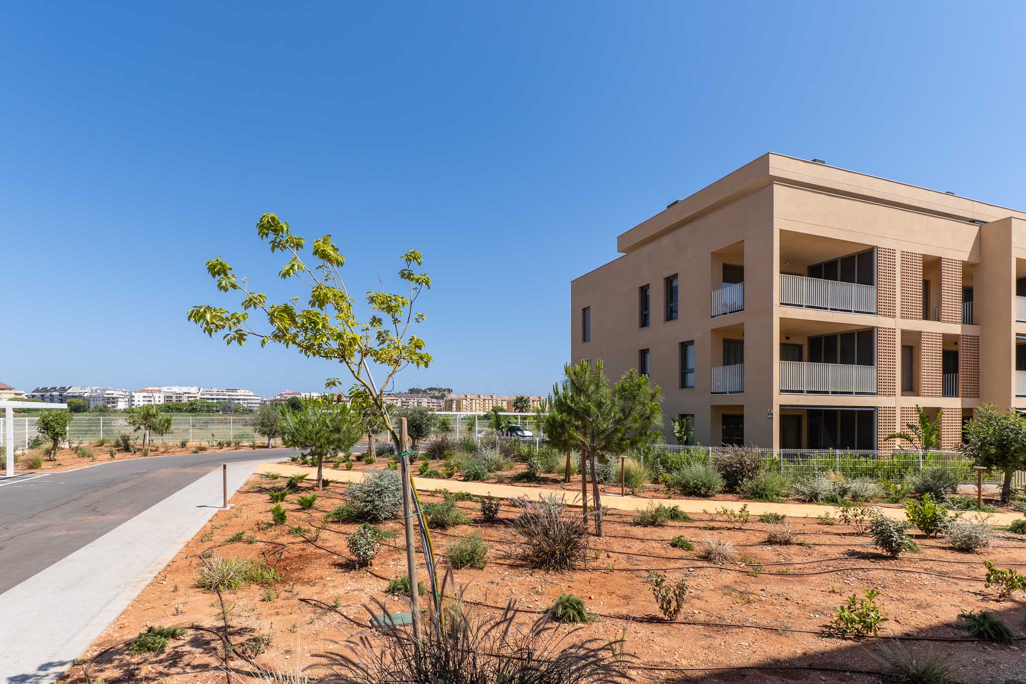 Exterior del moderno edificio residencial con fachada color arena y balcones de barandilla blanca. El entorno cuenta con paisajismo mediterráneo joven y camino asfaltado.