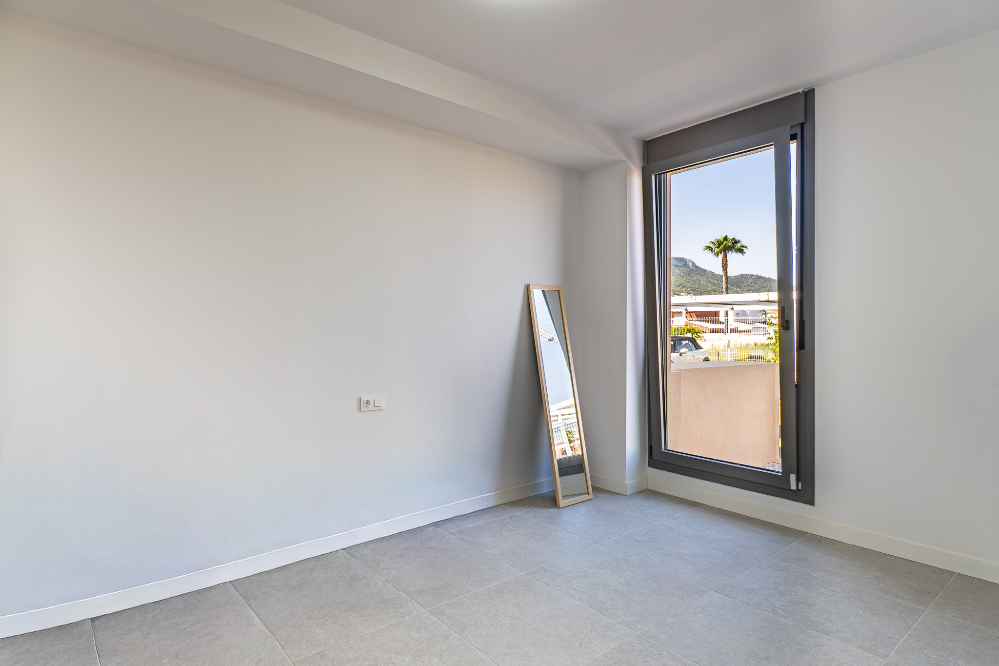 Habitación vacía con paredes blancas y suelo de gres gris, lista para personalizar. Gran ventanal con vistas a una palmera y montañas, aportando abundante luz natural.