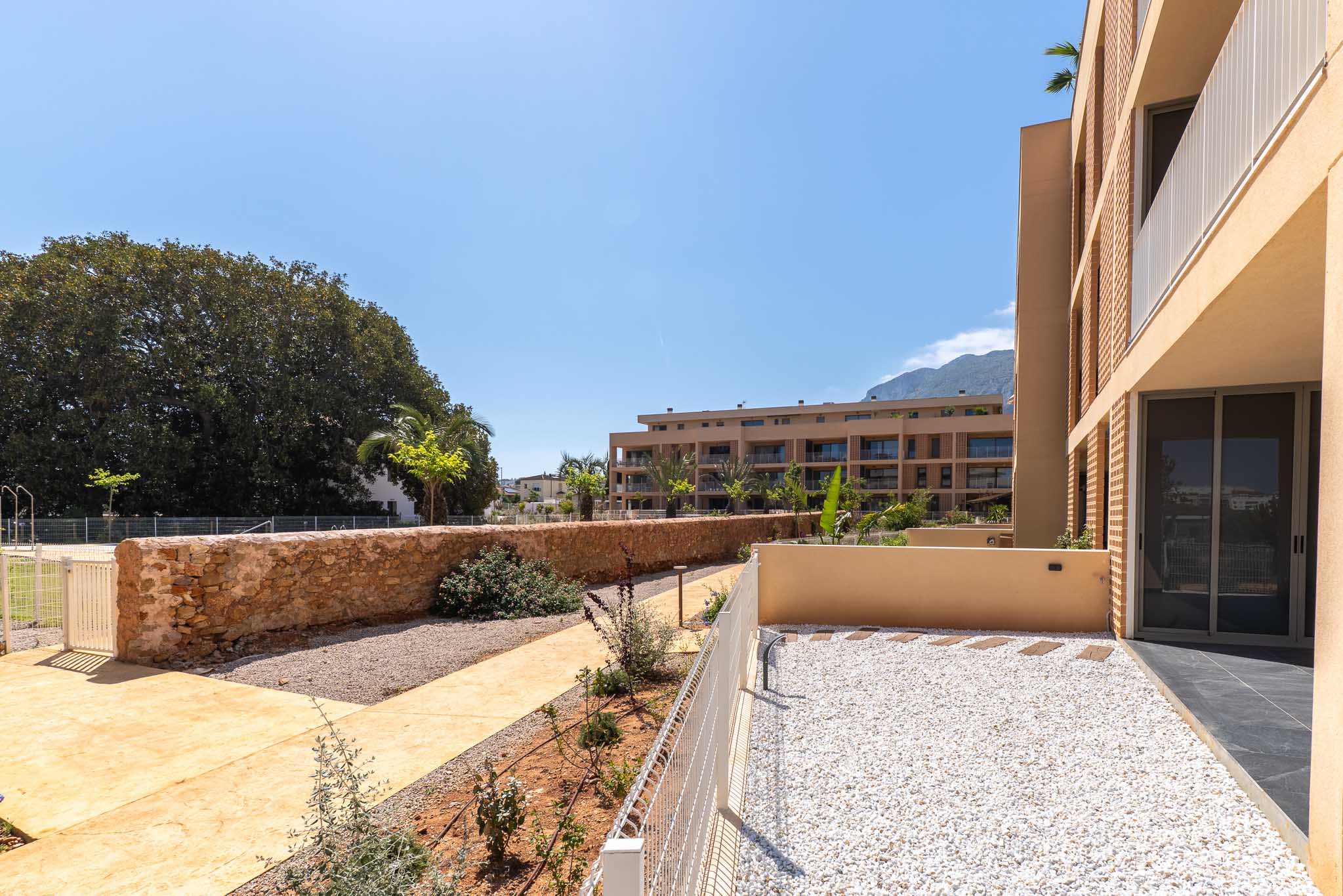 Jardín privado en planta baja con grava blanca, delimitado por una valla baja y una pared de piedra. Se aprecian senderos y vegetación joven, con vistas a edificios modernos y montañas.