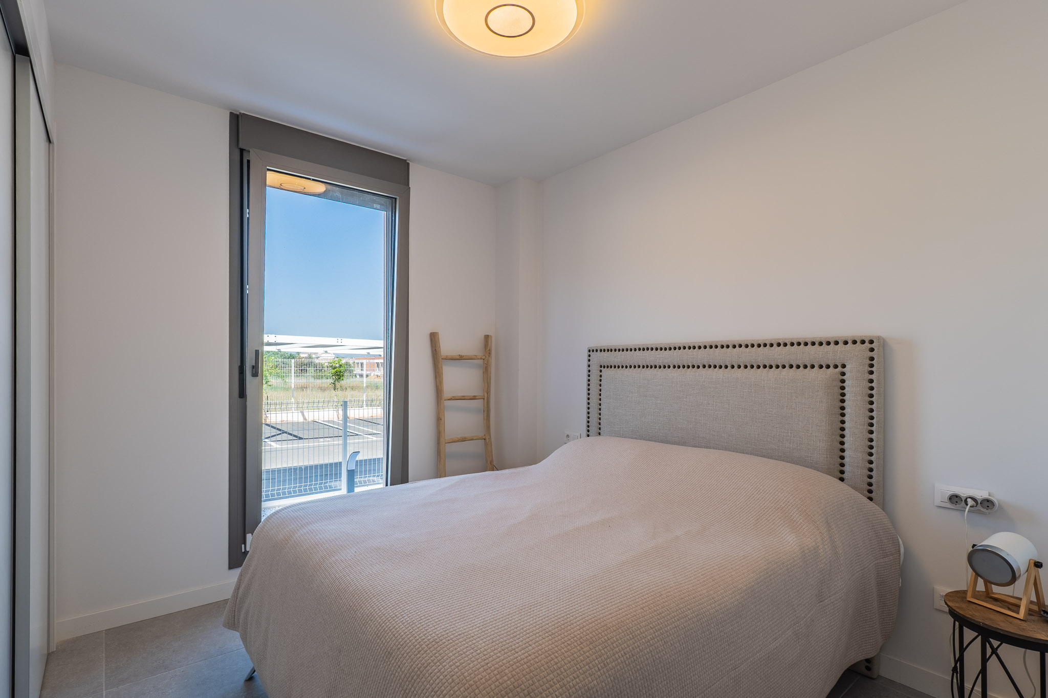 Luminosa habitación con cama, cabecero tapizado con tachuelas y armario empotrado. Gran ventanal con acceso al exterior y suelo de gres.