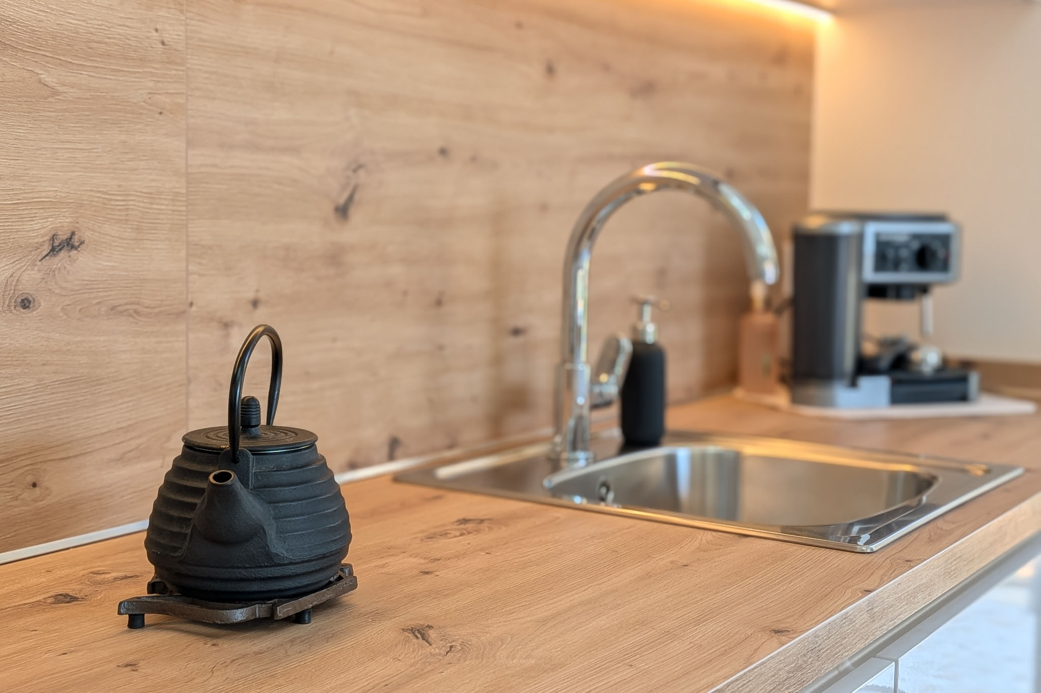Moderna encimera de cocina con acabado de madera clara y salpicadero a juego. Fregadero de acero inoxidable, grifo cromado y luz LED bajo mueble. Tetera de hierro fundido en primer plano.