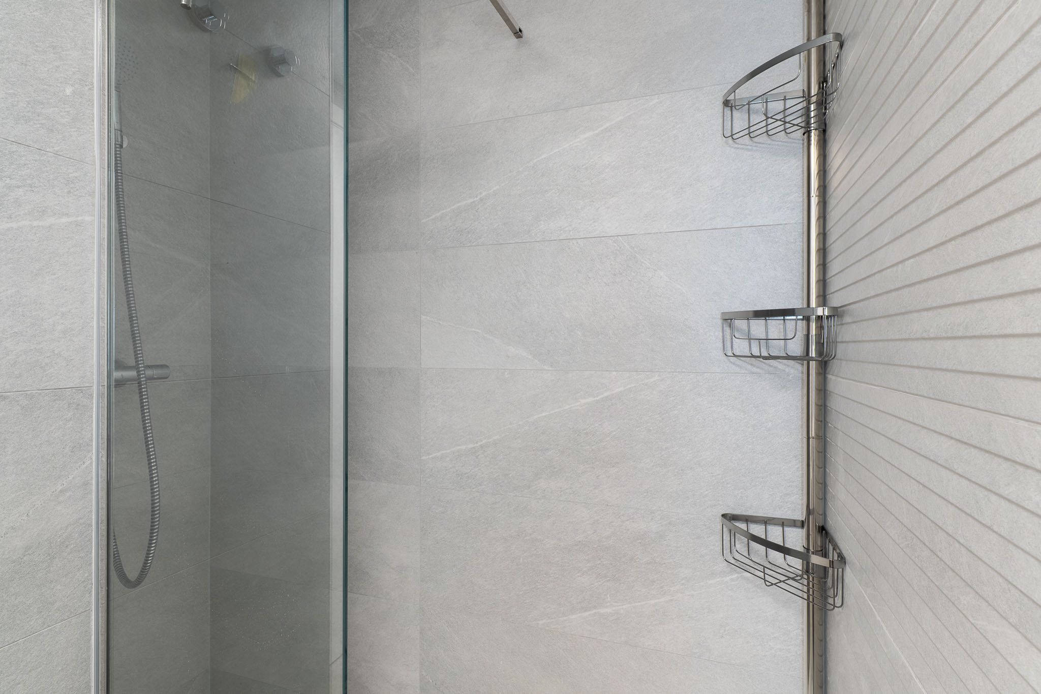 Moderna zona de ducha con azulejos grises de gran formato, una pared de acento texturizada, mampara de cristal y una práctica estantería esquinera cromada.