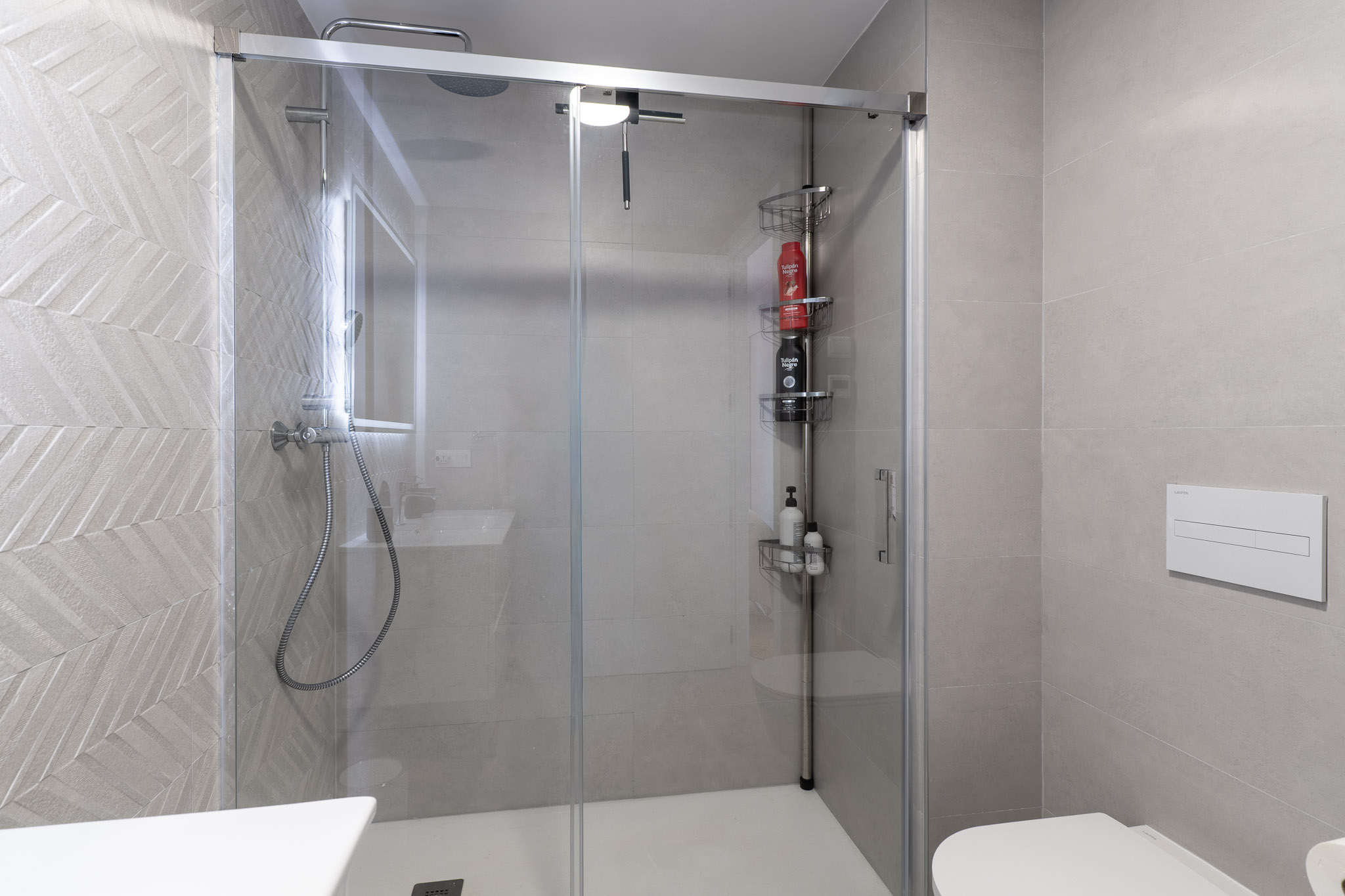 Moderno baño con cabina de ducha de cristal y plato blanco. Destacan los azulejos grises con patrón de espiga, ducha de lluvia y un inodoro suspendido.