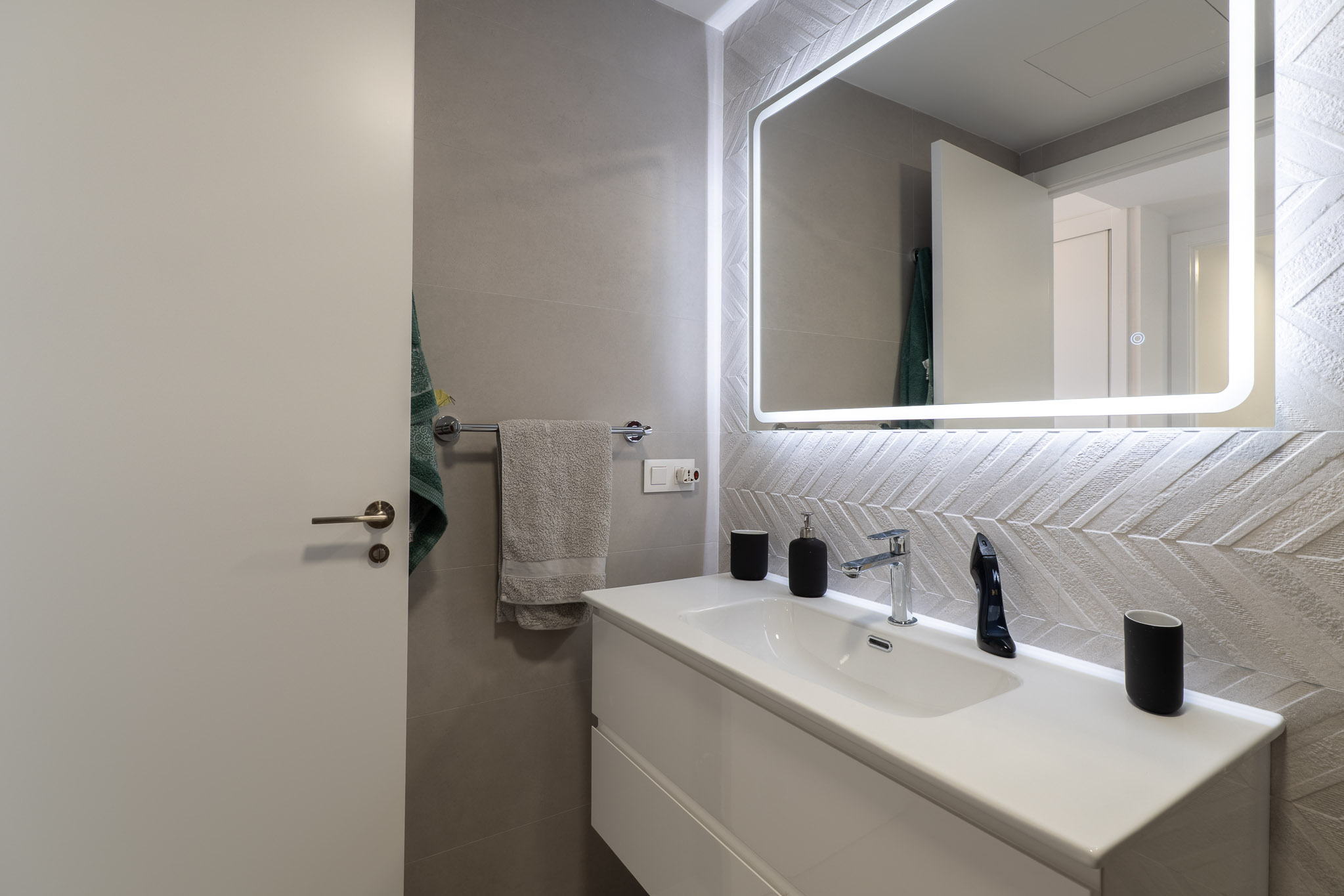 Moderno baño con lavabo flotante blanco, grifería cromada y espejo retroiluminado. Destacan los azulejos texturizados con patrón de espiga en la pared.