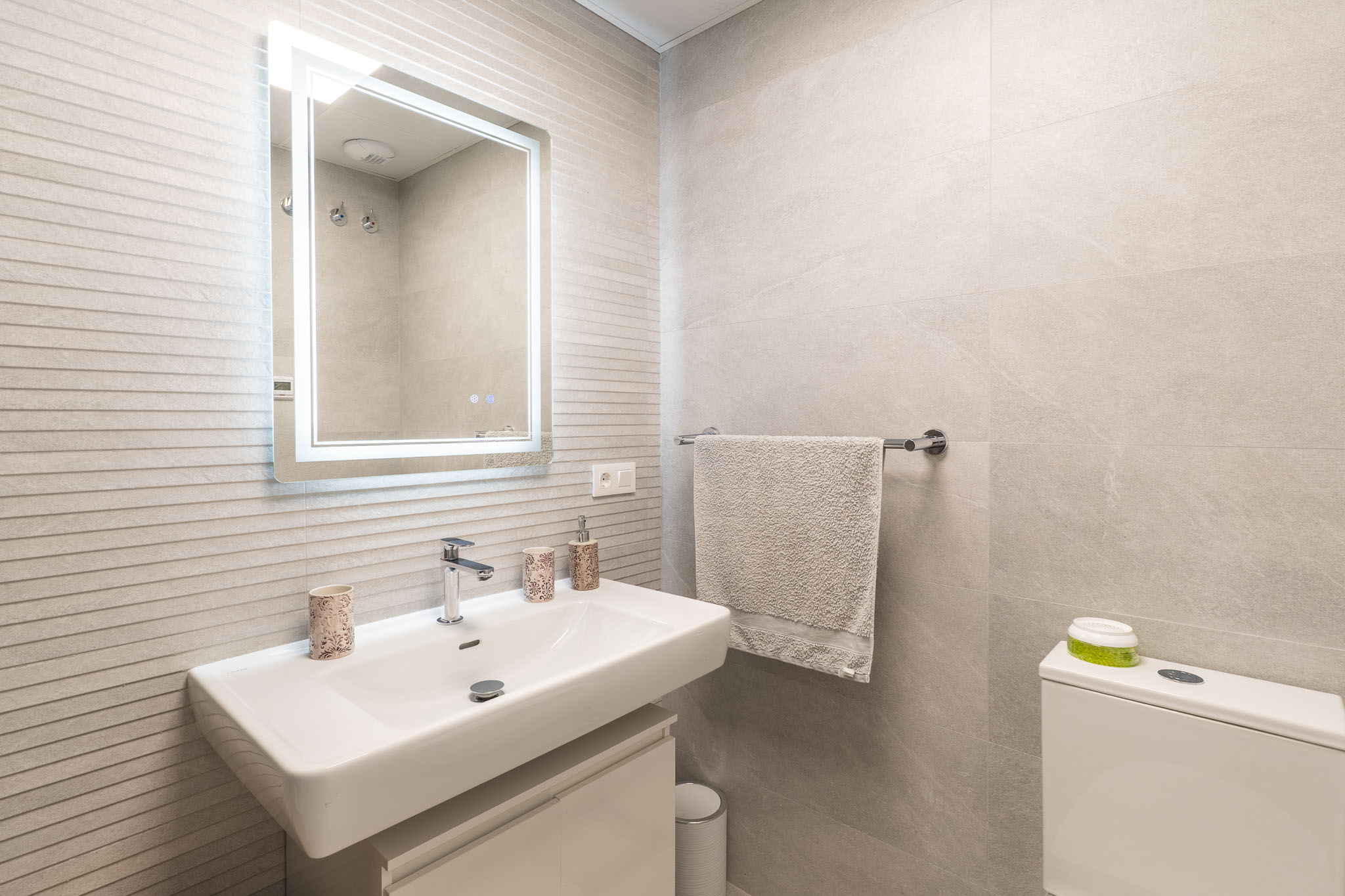 Moderno tocador de baño con lavabo rectangular blanco, grifo mural, espejo iluminado y azulejos de pared grises texturizados.