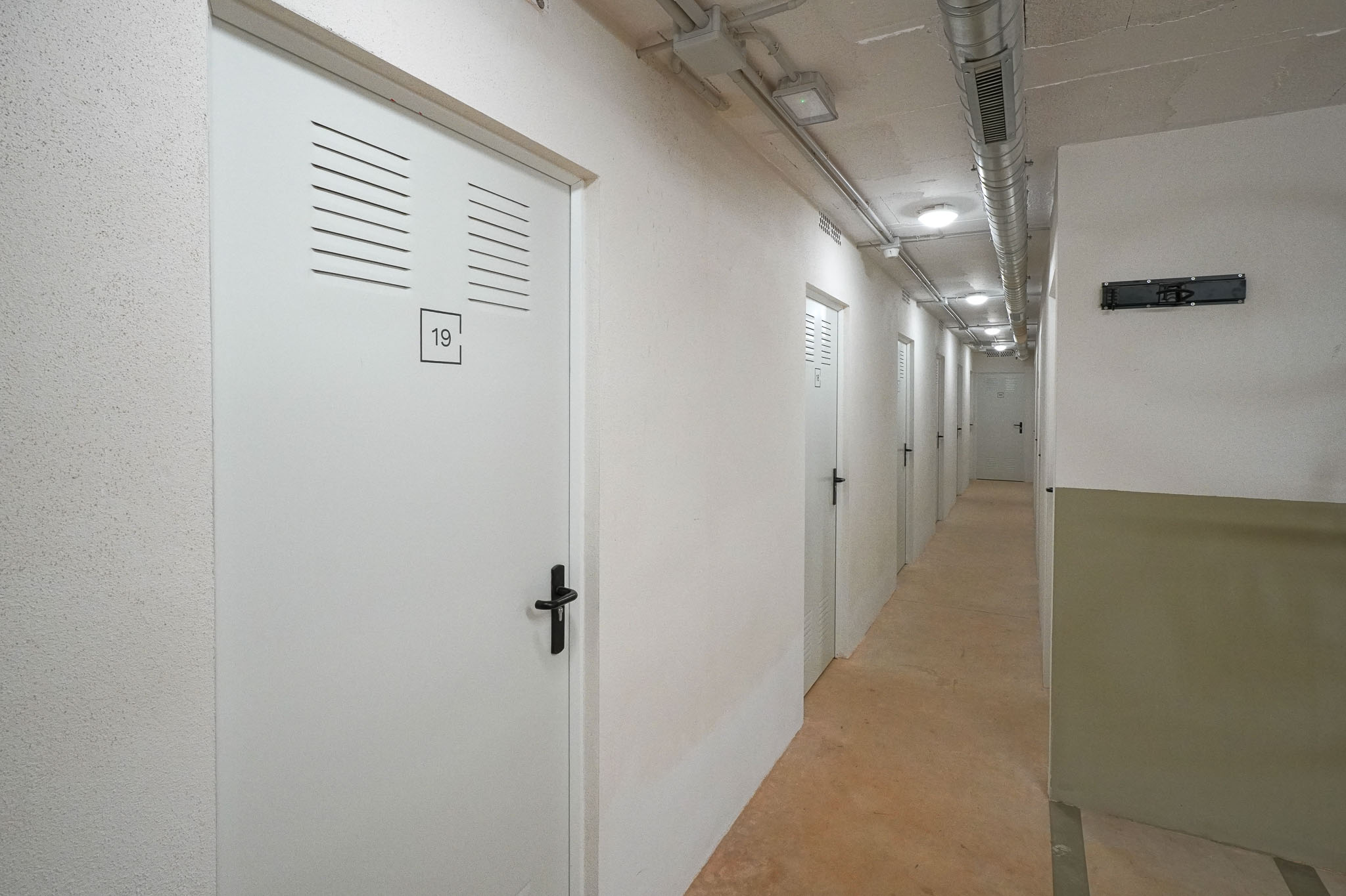 Pasillo de trasteros con puertas blancas numeradas y rejillas de ventilación. El techo muestra conductos de ventilación expuestos e iluminación empotrada, sobre un suelo de hormigón.