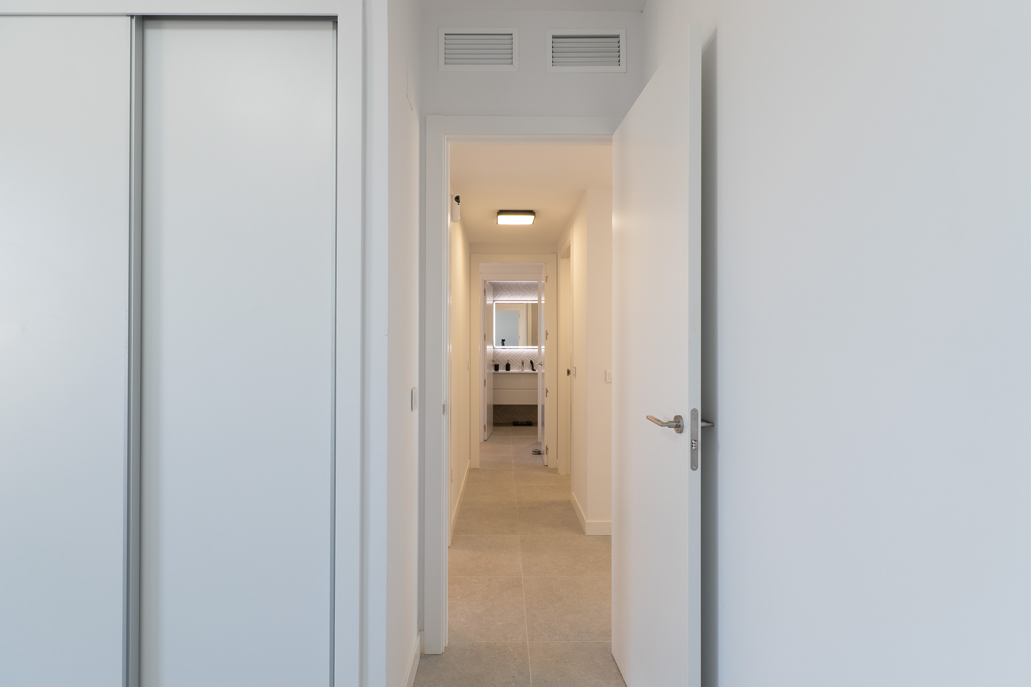 Pasillo luminoso con armario empotrado blanco y puerta abierta. El suelo de baldosas claras conduce a un baño moderno con espejo retroiluminado y mueble de diseño. Rejillas de ventilación en el techo.