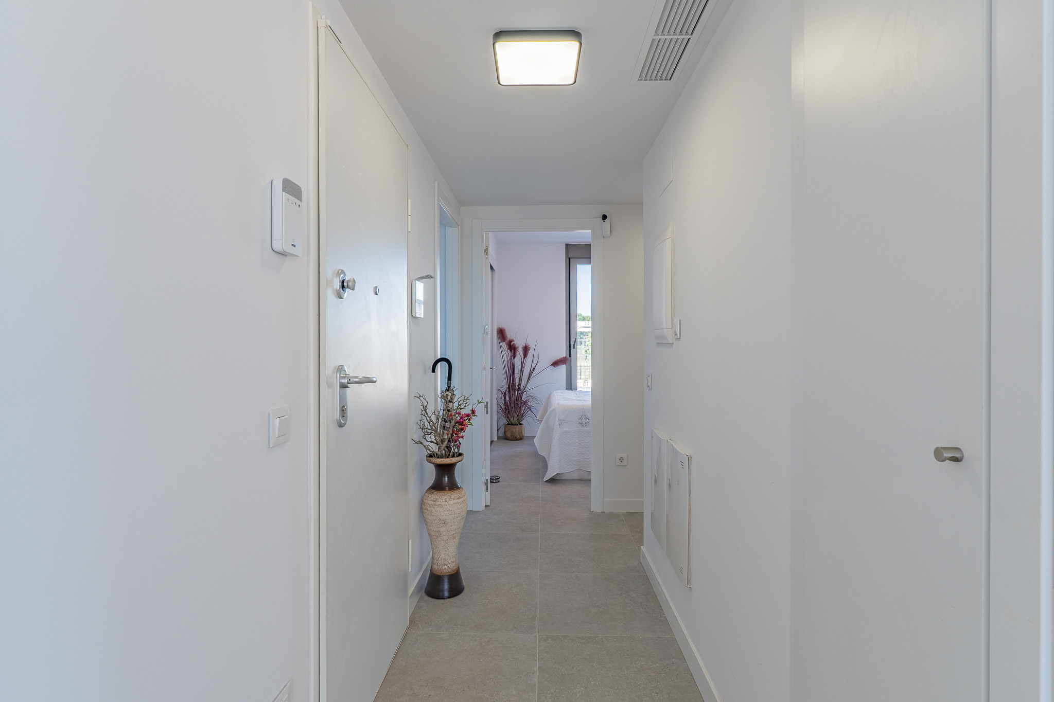 Pasillo moderno y luminoso con suelo de gres claro y paredes blancas. Incluye panel de seguridad, iluminación de techo contemporánea y vista a un dormitorio.