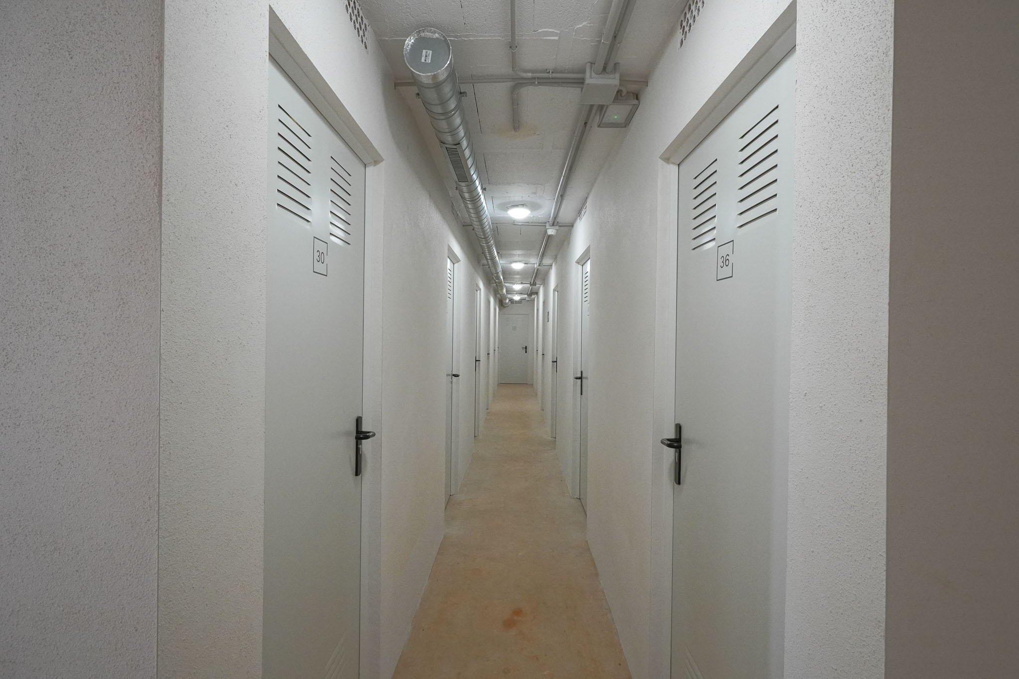 Pasillo utilitario a trasteros individuales, con puertas blancas numeradas y rejillas de ventilación. Techo de hormigón visto con conductos industriales e iluminación empotrada, junto a paredes texturizadas.