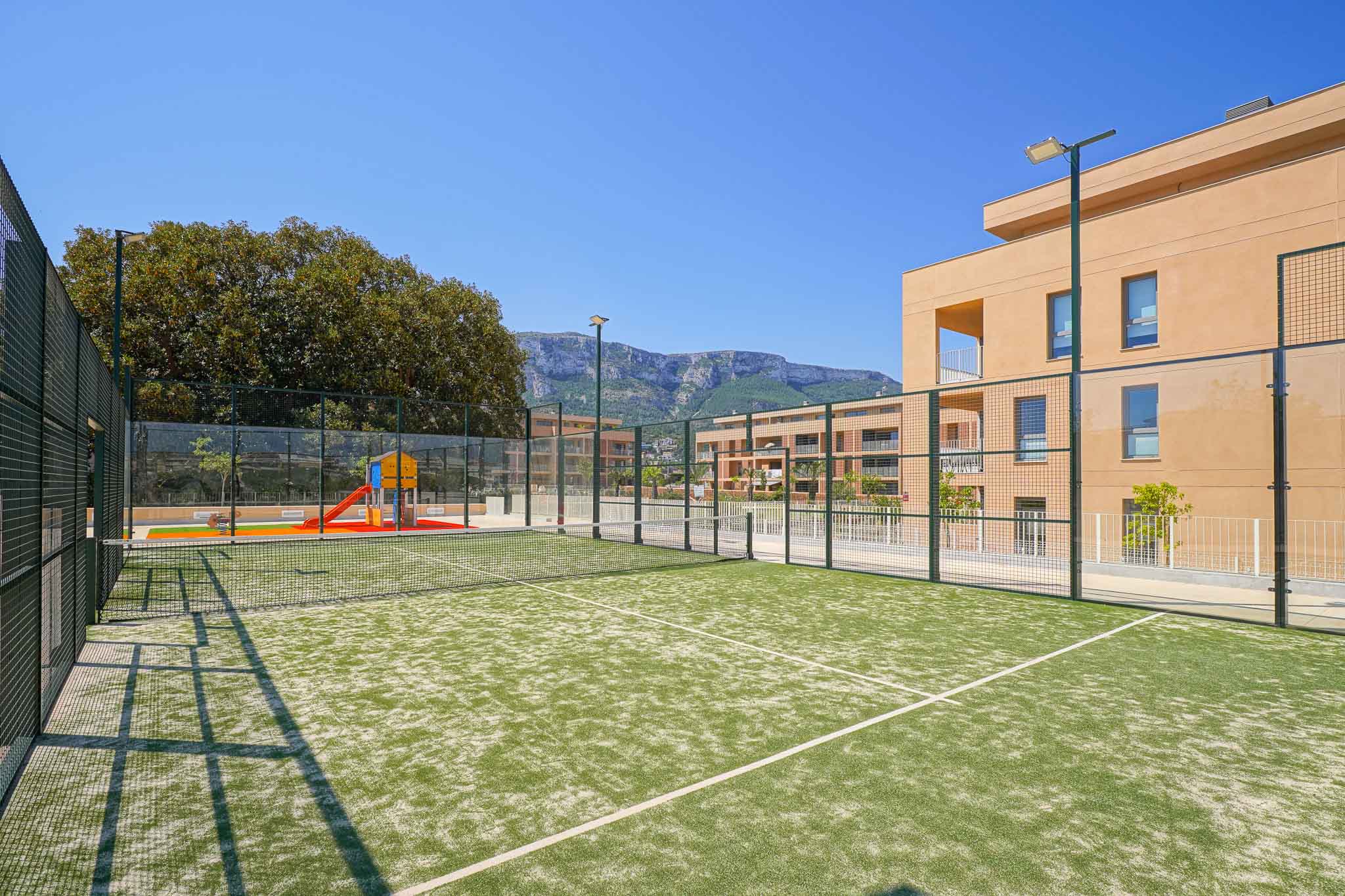 Pista de pádel de césped artificial verde, con vallas y postes de iluminación, junto a un parque infantil y edificios residenciales modernos con vistas a la montaña.