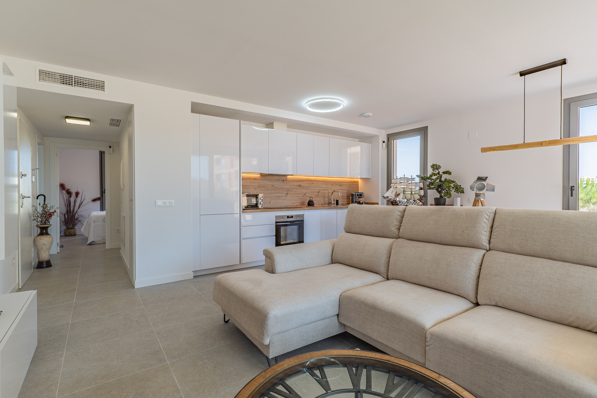 Salón-cocina diáfano con sofá seccional beige, cocina moderna de armarios blancos y encimera de madera clara. Suelo de gres y acceso a exterior.