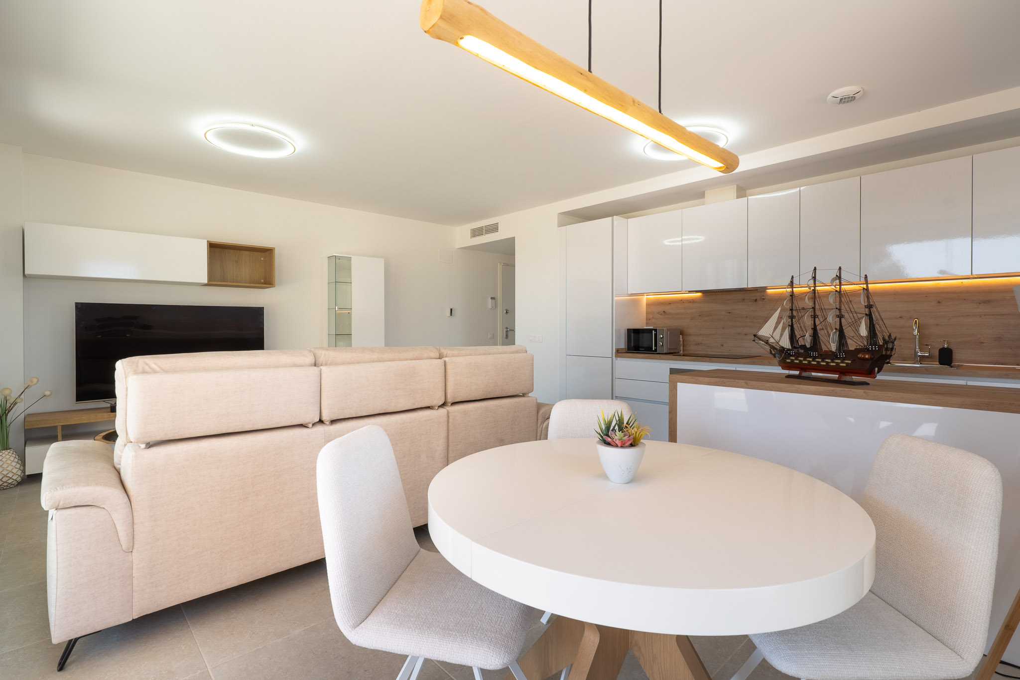 Salón-comedor de planta abierta con cocina integrada. Destaca el sofá beige, mesa ovalada blanca y cocina moderna con muebles lacados blancos, salpicadero de madera y luz ambiental.