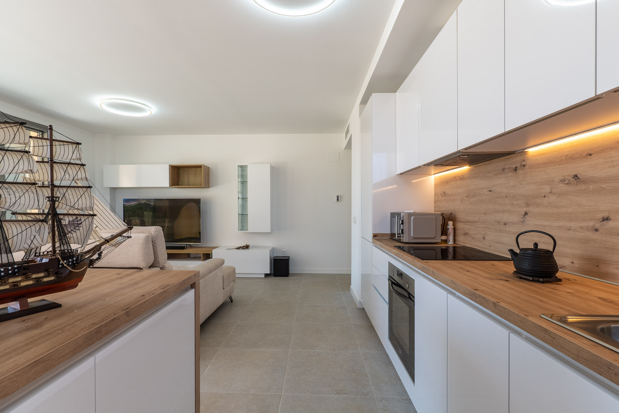 Salón-comedor diáfano con cocina abierta de diseño moderno. Destacan los muebles blancos lacados, encimeras de efecto madera clara y electrodomésticos integrados, complementados con iluminación LED y suelo cerámico gris.