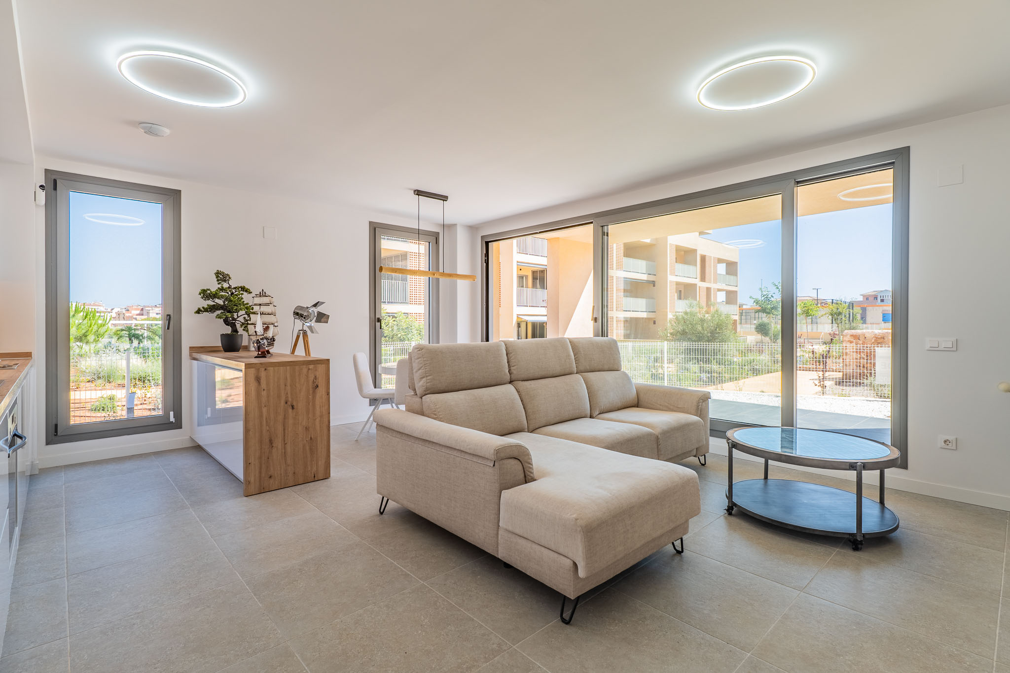 Salón-comedor diáfano con sofá seccional beige y mesa de centro ovalada. Grandes ventanales conectan con el exterior, ofreciendo luz natural y vistas residenciales. Cocina abierta con barra de madera.