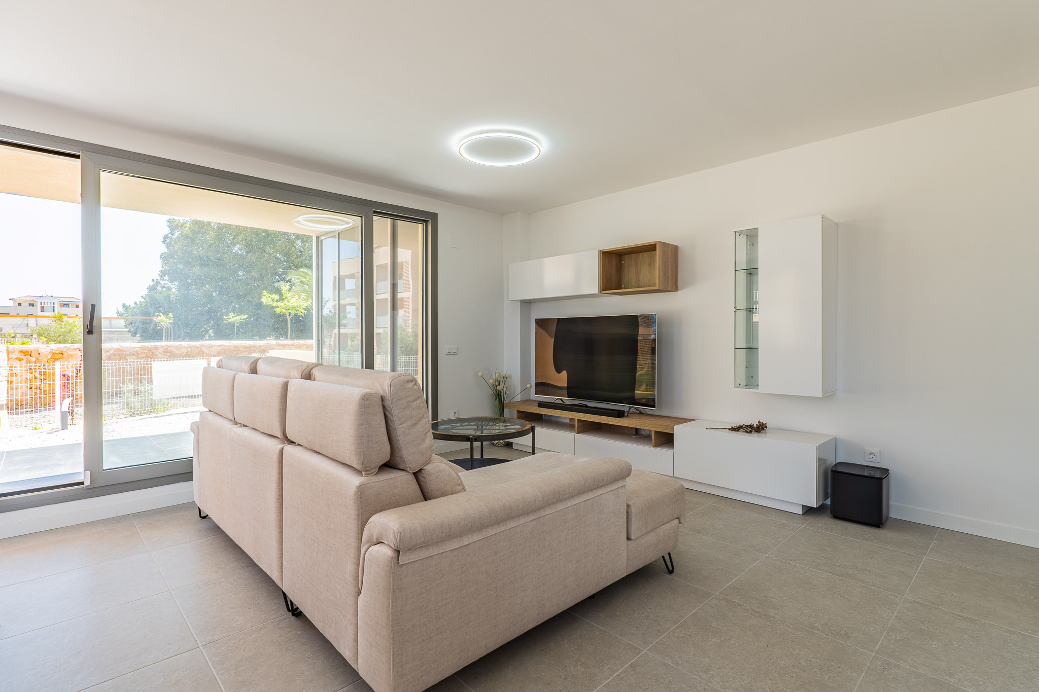 Salón luminoso con sofá seccional beige, suelo de gres claro y amplios ventanales correderos que dan al exterior. Mueble de TV moderno con estantes blancos y de madera.